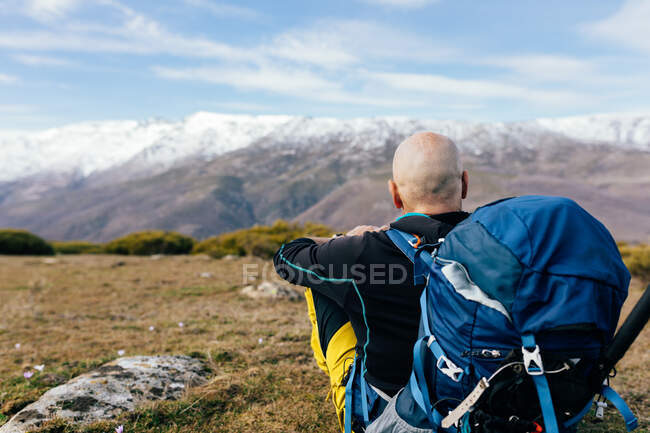 Rückenansicht eines nicht wiederzuerkennenden erwachsenen aktiven männlichen Bergsteigers in Aktivkleidung mit Rucksack, der sich auf einem grasbewachsenen Berggipfel ausruht und die malerische Aussicht auf die verschneite Bergkette genießt — Stockfoto
