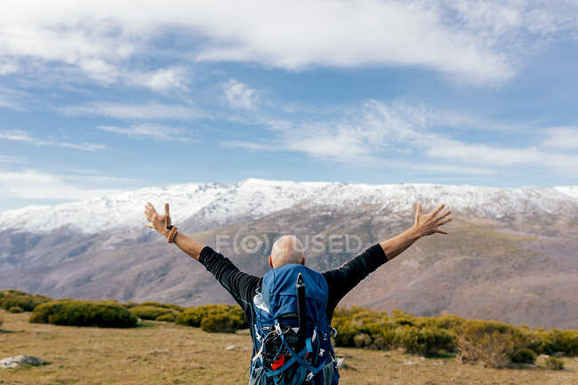 Обратный вид на неузнаваемого активного лысого альпиниста в красочной одежде, поднимающего руки и наслаждающегося свободой, стоя на снежной вершине горы против голубого облачного неба в солнечный день в высокогорье — стоковое фото