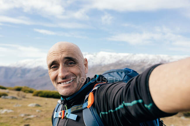 Homme adulte joyeux grimpeur en vêtements de sport regardant la caméra tout en prenant selfie contre majestueux pics rocheux enneigés dans la journée ensoleillée — Photo de stock