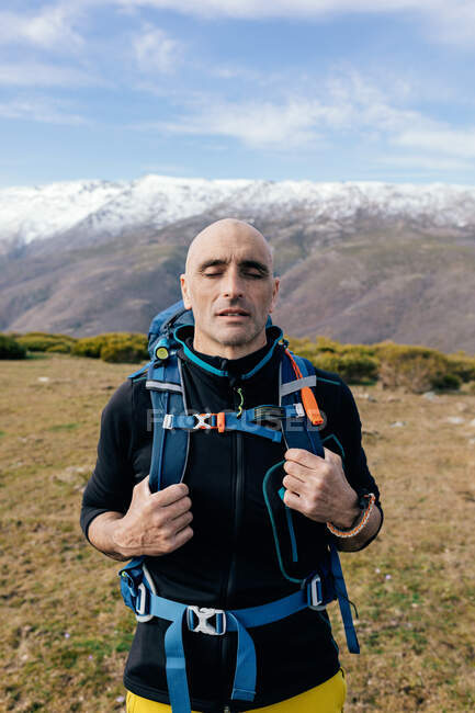 Adult bald active male mountaineer with eyes closed in activewear with backpack getting rest on mountain top and enjoying picturesque view of snow covered range — Stock Photo