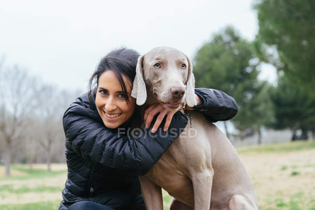Cheerful female owner embracing loyal purebred Weimaraner dog while sitting together on border on grassy meadow in park — Stock Photo