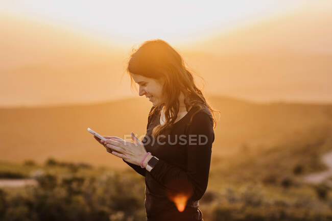 Вид сбоку восхищенной женщины, просматривающей мобильный телефон, стоя на холме на фоне горы под закатным небом — стоковое фото