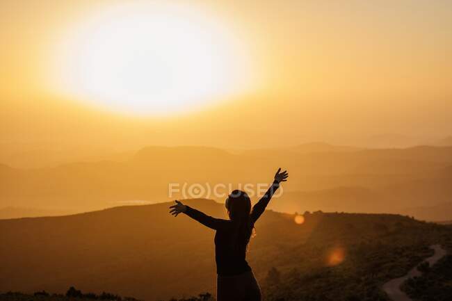 Back view of unrecognizable female standing with raised arms on hill and enjoying freedom while admiring mountainous scenery at sunset — Stock Photo