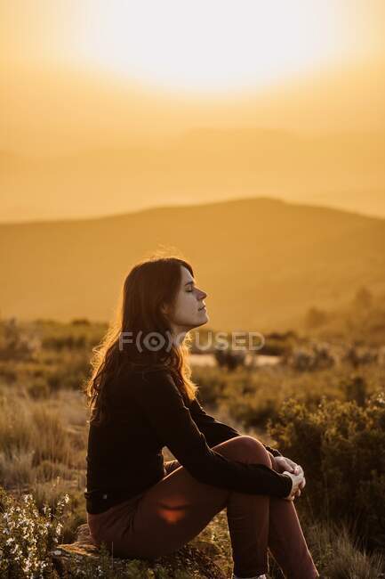 Seitenansicht einer friedlichen Reisenden, die mit geschlossenen Augen auf einem Hügel sitzt und die Natur im Hochland bei Sonnenuntergang genießt — Stockfoto