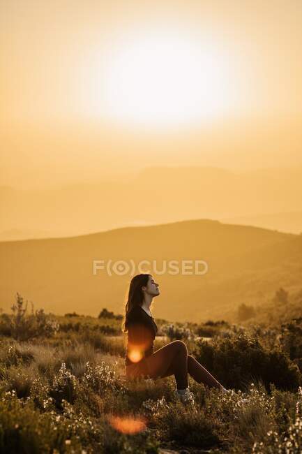 Vista lateral de la tranquila viajera sentada en la colina con los ojos cerrados y disfrutando de la naturaleza en las tierras altas al atardecer - foto de stock