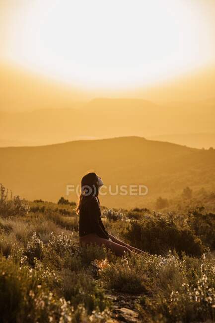Vista laterale di tranquillo viaggiatore femminile seduto sulla collina con gli occhi chiusi e godersi la natura negli altopiani al tramonto — Foto stock