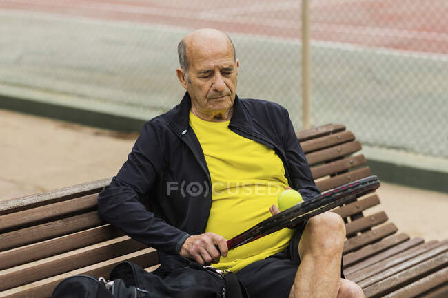 Пожилой спортсмен с ракеткой и мячом сидит на скамейке на корте перед теннисными тренировками — стоковое фото