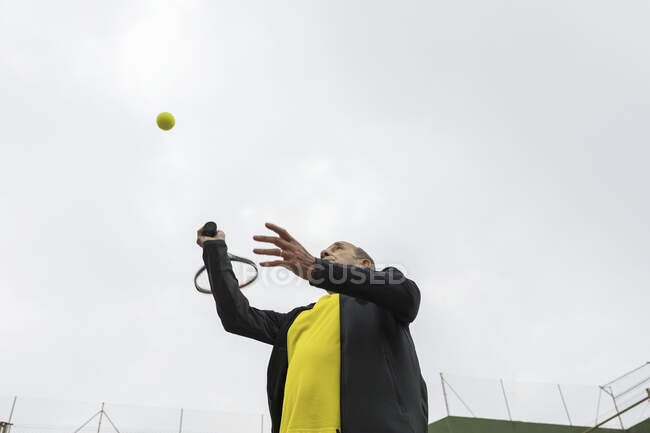 Пожилой спортсмен бьет по мячу ракеткой во время игры в теннис на зеленой площадке в пасмурный день — стоковое фото