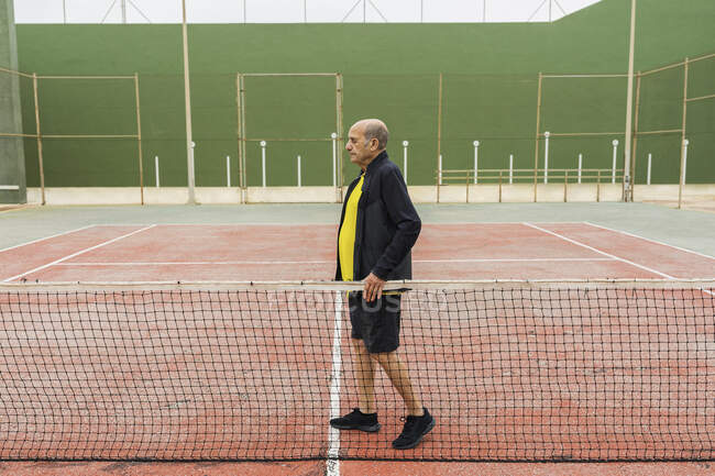 Vista lateral homem idoso caminhando perto da rede durante o treinamento na quadra de tênis — Fotografia de Stock