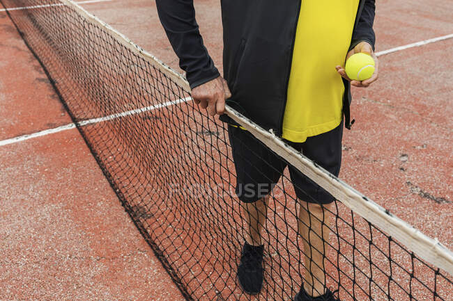 De arriba anónimo anciano con pelota de pie cerca de la red durante el entrenamiento en la cancha de tenis - foto de stock