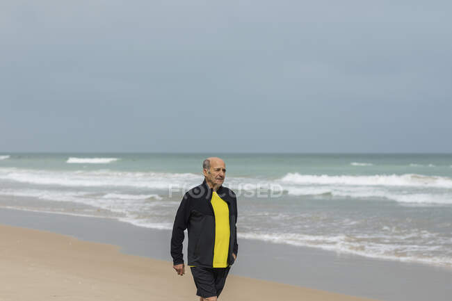 Anciano deportista caminando en la playa cerca del mar ondeando durante el entrenamiento de fitness en verano - foto de stock