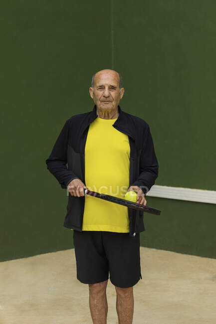Elderly sportsman with tennis ball and racket looking at camera while standing against green wall during workout in gym — Stock Photo