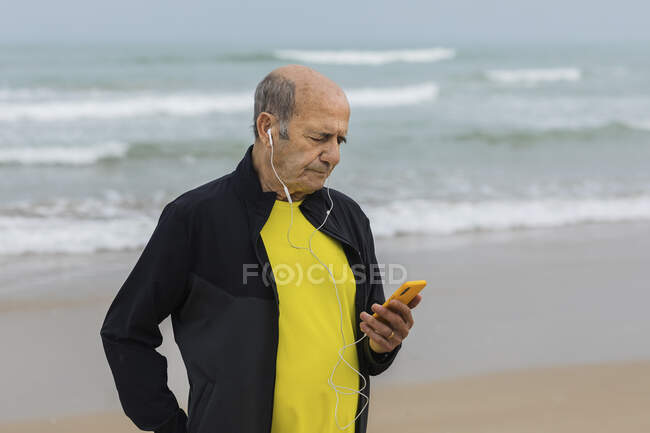 Aged male athlete listening to music in earphones and using smartphone during workout on beach near waving sea — Stock Photo