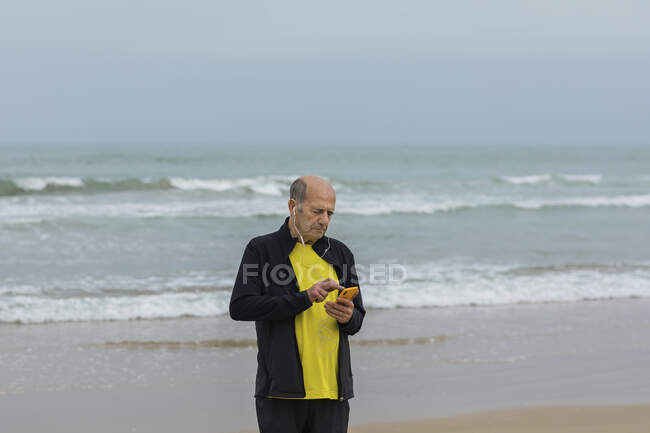 Atleta do sexo masculino envelhecido ouvindo música em fones de ouvido e usando smartphone durante o treino na praia perto do mar ondulado — Fotografia de Stock