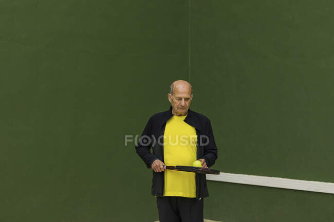Elderly sportsman with tennis ball and racket looking down while standing against green wall during workout in gym — Stock Photo