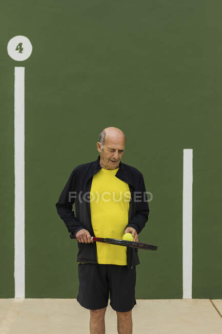 Літній спортсмен з тенісним м'ячем і ракеткою, дивлячись вниз, стоячи на зеленій стіні під час тренувань у спортзалі — стокове фото