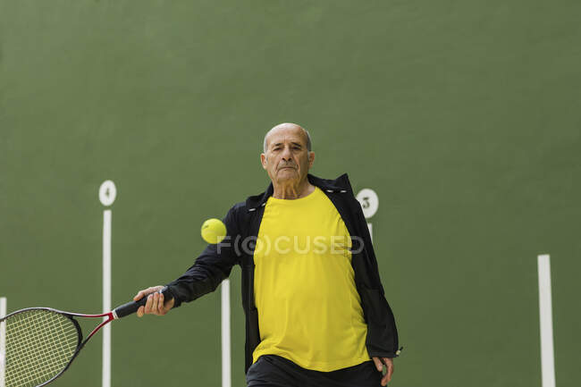 Пожилой спортсмен бьет по мячу ракеткой, играя в теннис против зеленой стены в спортзале — стоковое фото
