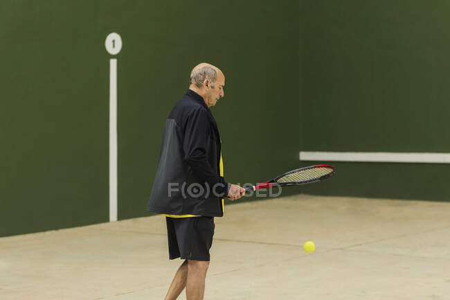 Elderly male athlete hitting ball with racket while playing tennis against green wall in gym — Stock Photo