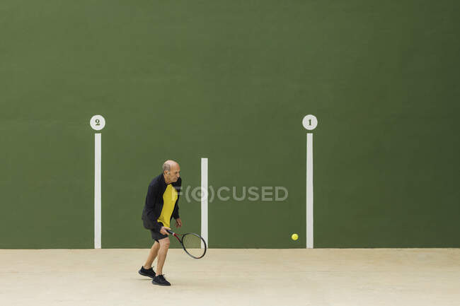 Elderly male athlete hitting ball with racket while playing tennis against green wall in gym — Stock Photo