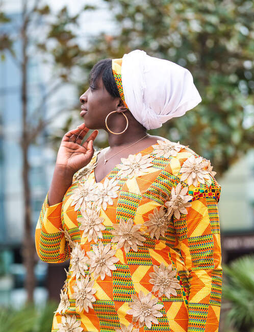 Вид сбоку на молодую африканскую женщину в яркой одежде с цветочными вставками, смотрящую в сторону стоя в городе — стоковое фото