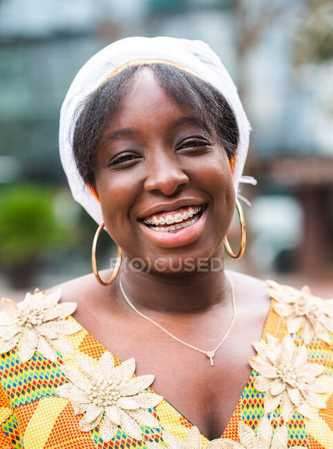 Cheerful young African female in bright wear with floral ornament looking at camera standing in the city — Stock Photo