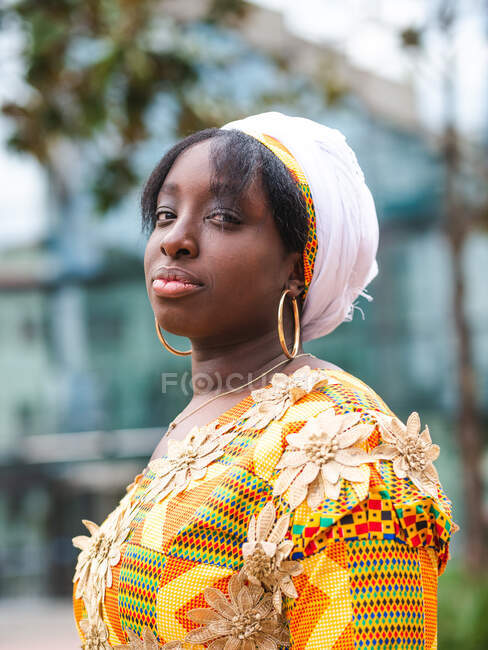 Vista laterale della giovane donna africana in abito luminoso con ornamento floreale guardando la fotocamera in piedi in città — Foto stock