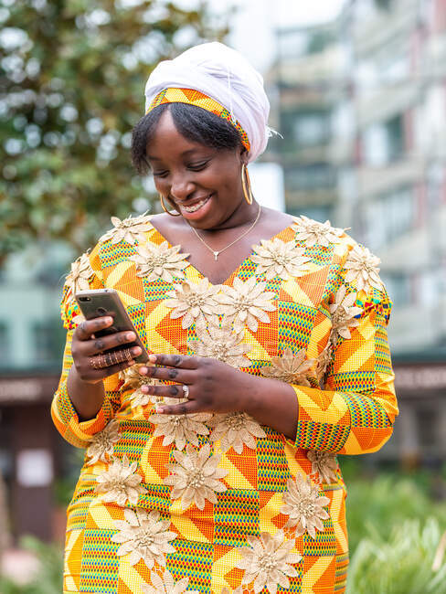 Dal basso di sorridente femmina nera in abiti ornamentali navigando sul cellulare mentre in piedi in città — Foto stock