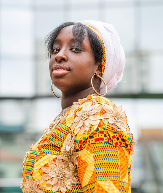 Vista lateral da jovem fêmea africana em desgaste brilhante com ornamento floral olhando para a câmera em pé na cidade — Fotografia de Stock