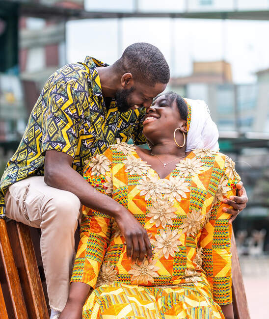Веселая африканская женщина в традиционной одежде сидит рядом с небритым парнем в декоративной одежде на городской скамейке — стоковое фото