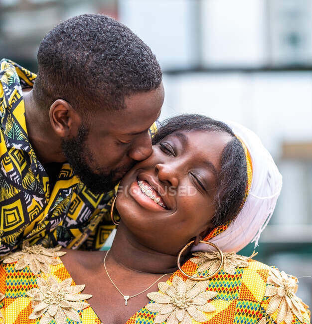 Cheerful African man in traditional wear kissing cheek smiling girlfriend in ornamental clothes on city bench — Stock Photo