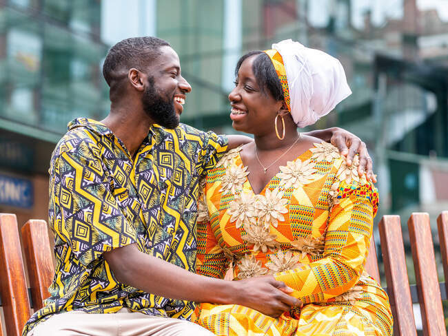 Cheerful African woman in traditional wear sitting close to unshaven boyfriend in ornamental clothes on city bench — Stock Photo