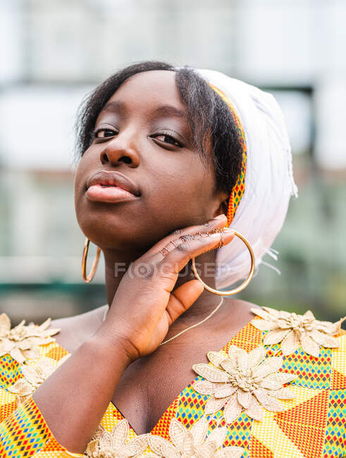Young African female in bright wear with floral ornament looking at camera standing in the city — Stock Photo