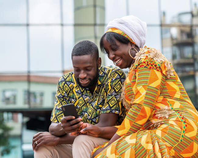 Cheerful African American man with content female beloved in traditional wear with ornament surfing internet on cellphone in town — Stock Photo