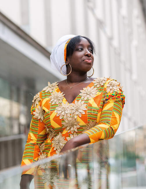 Joven mujer africana en ropa brillante con adorno floral mirando hacia otro lado cerca de la valla de vidrio en la ciudad - foto de stock