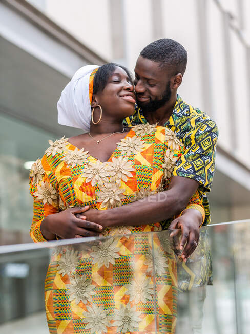 Ethnischer Mann mit geschlossenen Augen umarmt und küsst Partnerin in heller traditioneller Kleidung mit Ornament in der Stadt — Stockfoto