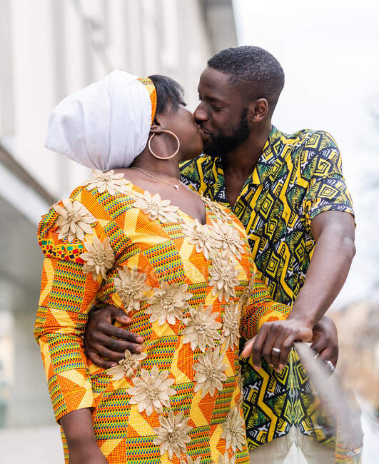 Homem étnico com olhos fechados abraçando e beijando parceiro feminino em roupas tradicionais brilhantes com ornamento na cidade — Fotografia de Stock