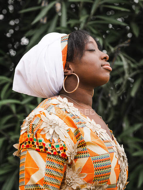 Vista lateral de una joven africana en pañuelo con los ojos cerrados contra la planta en verano - foto de stock