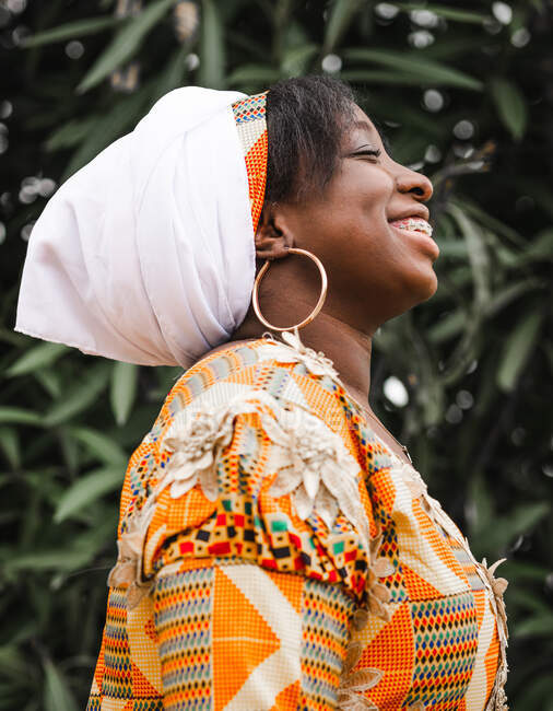 Vista lateral de la joven hembra africana alegre con tirantes en el pañuelo con los ojos cerrados contra la planta en verano - foto de stock