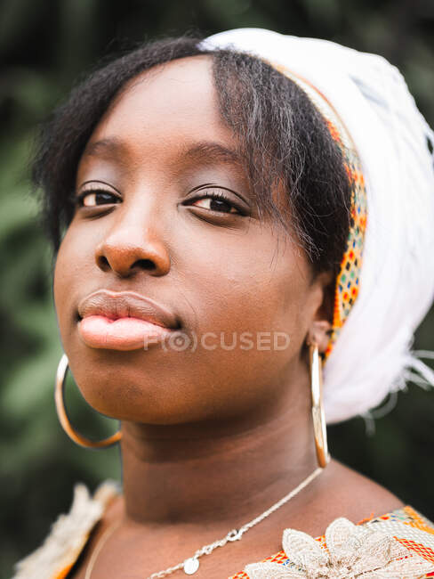 Vue latérale de la jeune femme africaine en mouchoir regardant caméra contre plante en été — Photo de stock