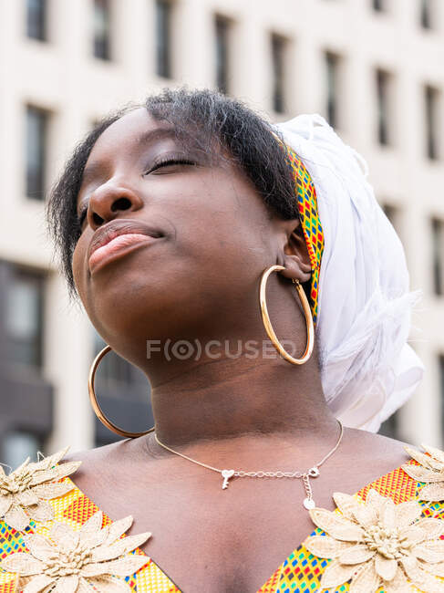 Jovem fêmea africana em desgaste brilhante com ornamento floral com os olhos fechados em pé na cidade — Fotografia de Stock
