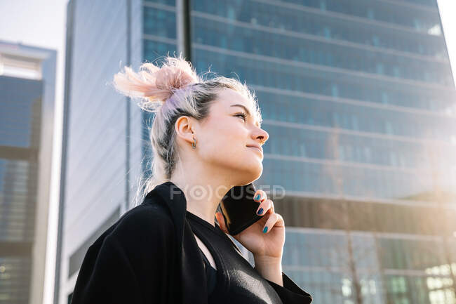Young cheerful female with hair bun talking on cellphone while looking away on city street in back lit — Stock Photo