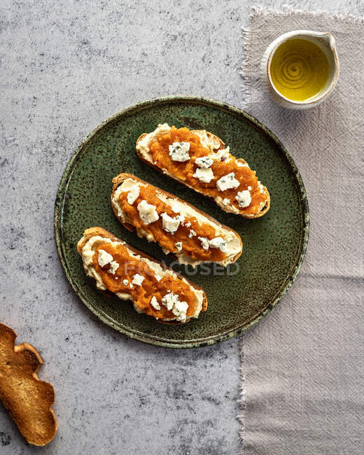 Вид сверху аппетитный свежие тосты с джемом и козьим сыром подается на тарелке рядом с кувшином с органическим оливковым маслом — стоковое фото