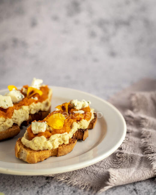 Свежеиспеченные тосты из тыквенного хлеба со сладким джемом и козьим сыром, украшенные лепестками желтых цветов и подаваемые на тарелке — стоковое фото