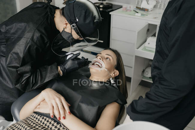 Angle élevé de patiente avec rétracteur et éjecteur de salive dans la bouche regardant le dentiste pendant la procédure dentaire en clinique — Photo de stock