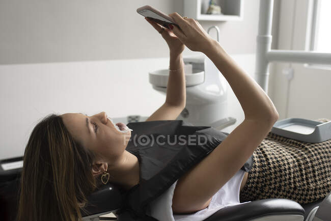 Vue latérale du patient féminin avec rétracteur dans la bouche couché sur une chaise dentaire et se faisant tirer dessus sur smartphone avant le traitement en clinique — Photo de stock