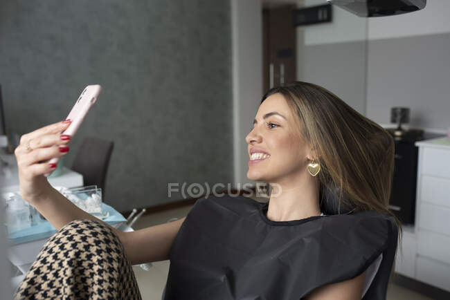 Paciente femenina con dientes blancos sentada en silla dental y autorretrato en smartphone tras tratamiento en clínica - foto de stock