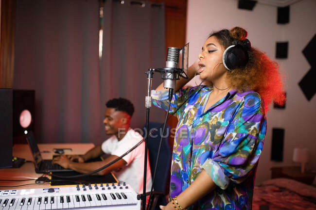 Боковой вид юной афроамериканки в наушниках, записывающей песню против улыбающегося музыкального продюсера в студии — стоковое фото