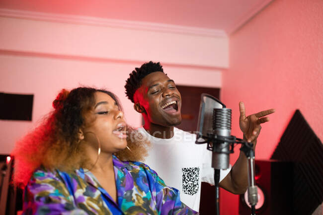 Jóvenes vocalistas afroamericanos con peinado afro y ojos cerrados cantando en micrófono profesional - foto de stock