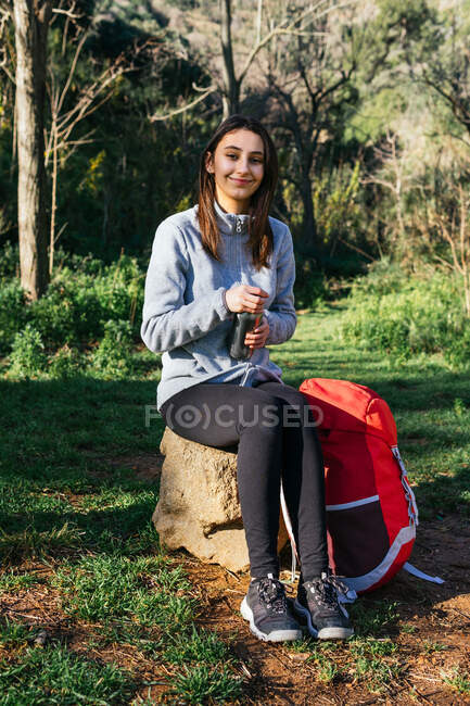 Fröhliche Wanderin mit Rucksack sitzt auf Baumstumpf im Wald und blickt in die Kamera — Stockfoto