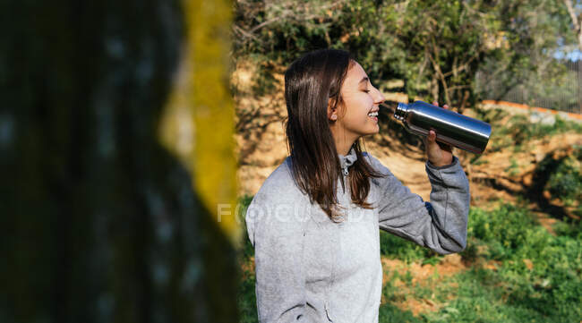 Seitenansicht einer Wanderin, die im Wald steht und beim Wandern frisches Wasser aus einer Metallflasche genießt — Stockfoto